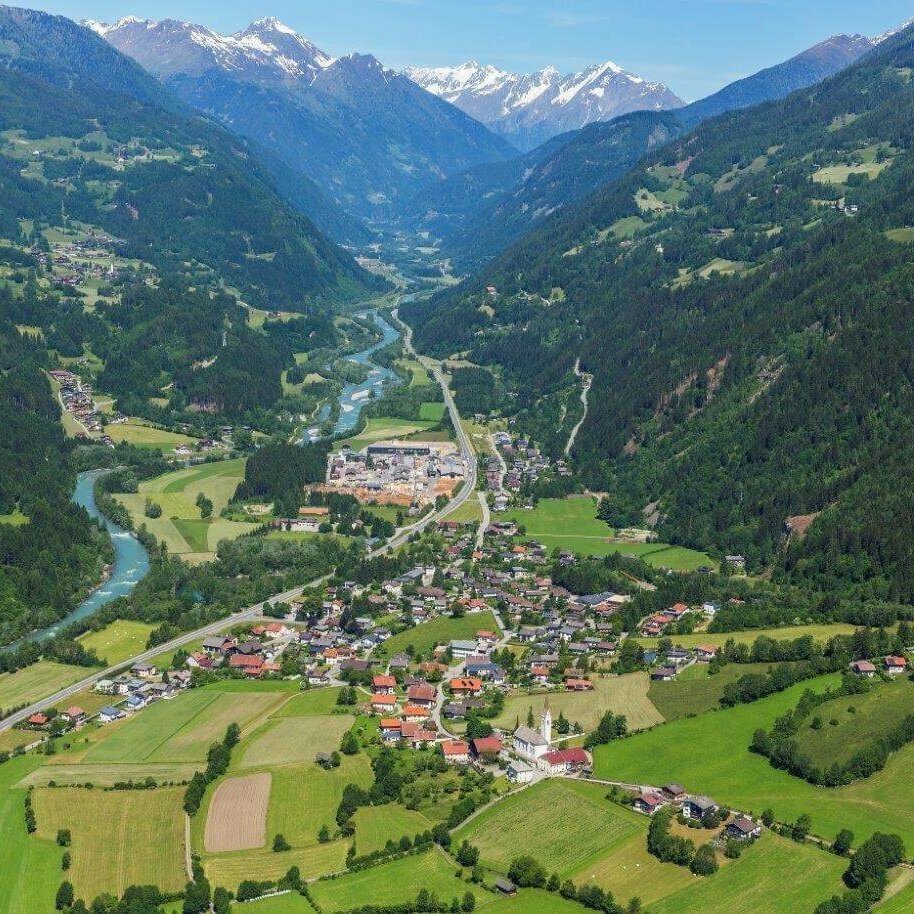 Ainet Luftaufnahme von Ainet im Sommer mit den grünen Wiesen und den Waldhängen. Im Hintergrund leuchten von den Bergen noch einige Schneefelder herunter.