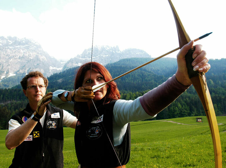 Bogensport Osttirol Ein Mann erklärt einer Frau die richtige Benützung eines Bogens. Die Frau hat den gespannten Bogen vor sich und zielt.