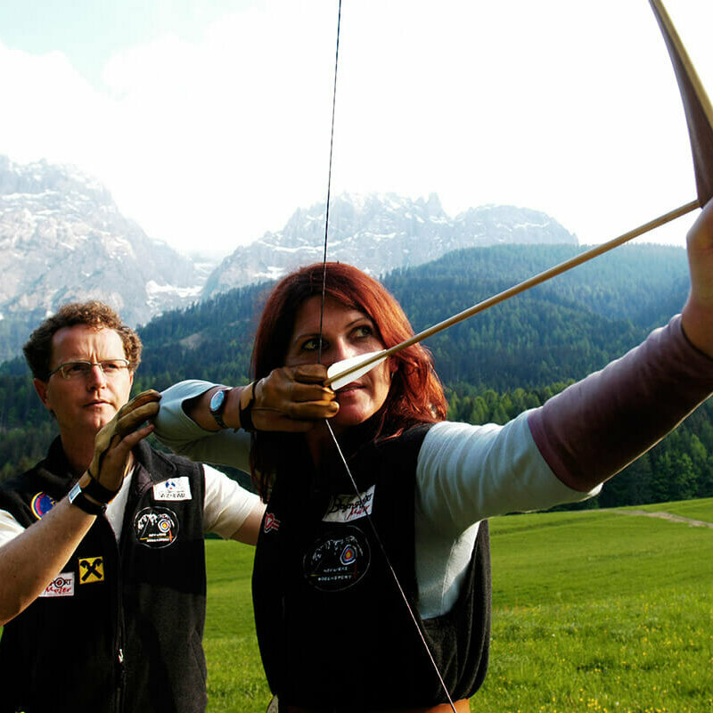 Bogensport Osttirol Ein Mann erklärt einer Frau die richtige Benützung eines Bogens. Die Frau hat den gespannten Bogen vor sich und zielt.