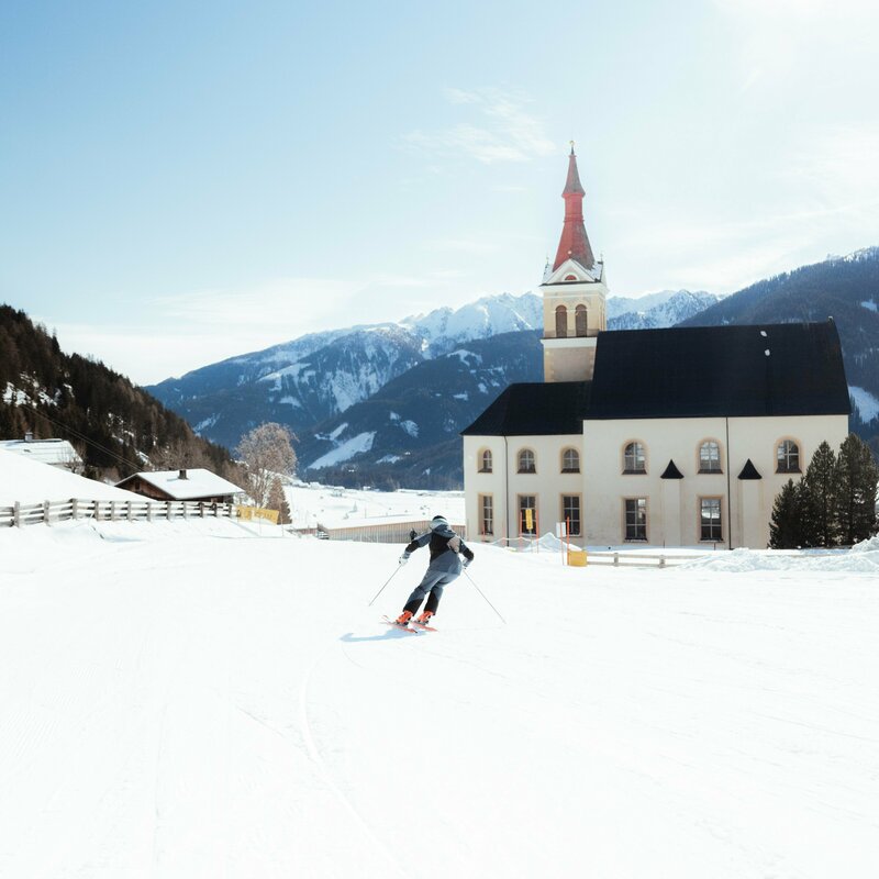 Ein Skifahrer zieht auf einer flachen Piste, die neben der Pfarrkirche vorbei läuft, bei herrlichem Winterwetter sein Schwünge sanft in den Schnee.