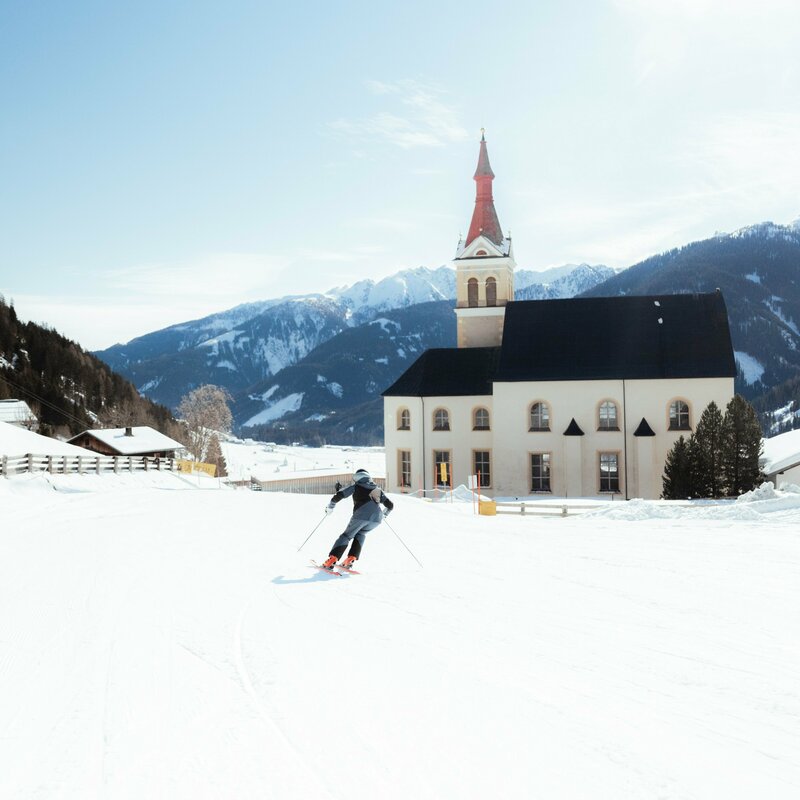 Ein Skifahrer zieht auf einer flachen Piste, die neben der Pfarrkirche vorbei läuft, bei herrlichem Winterwetter sein Schwünge sanft in den Schnee.