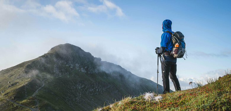 (Weit-)Wandern am Karnischen Höhenweg Schöne, frische Morgenstimmung - ein gut eingepackter Wanderer mit langer, dunkelgrauer Berghose, blauem Anorak mit übergezogener Kapuzze und blau-grauem Rucksack und angesteckter heller Trinkflasche blickt zu dem in einiger Entfernung liegenden Bergziel, das sich noch vom morgendlichen, leichten Nebel befreit.