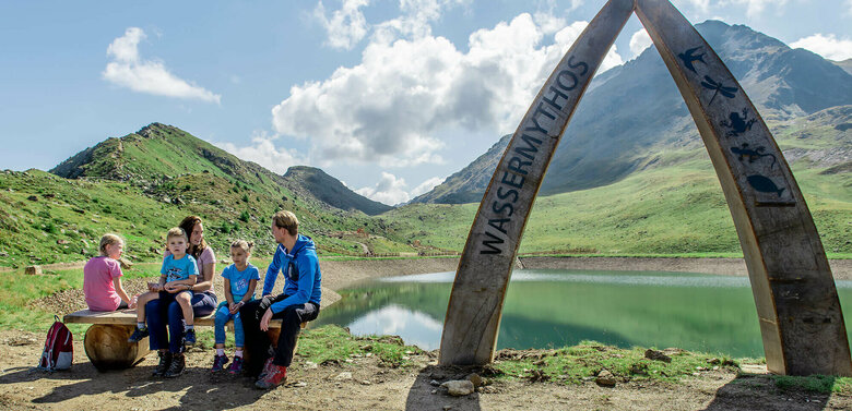 Eine Familie sitzt auf einer Bank am Ufer des Wassermythos Ochsenlacke im Skizentrum St. Jakob i. D..  Im Hintergrund sieht man die dazugehörigen Spielgeräte und das umliegende Bergpanorama, welches durch das schöne Wetter gut zur Geltung kommt.