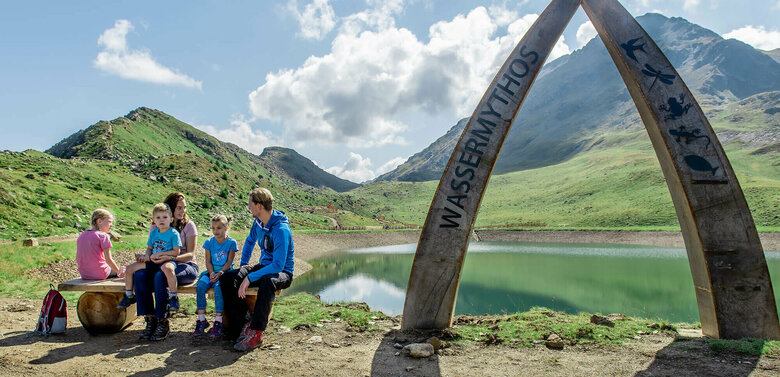 Wassermythos Ochsenlacke St. Jakob Brunnalm Eine Familie sitzt auf einer Bank am Ufer des Wassermythos Ochsenlacke im Skizentrum St. Jakob i. D.. Im Hintergrund sieht man die dazugehörigen Spielgeräte und das umliegende Bergpanorama, welches durch das schöne Wetter gut zur Geltung kommt.