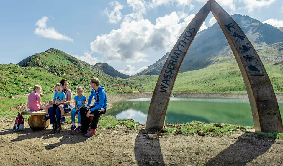 Eine Familie sitzt auf einer Bank am Ufer des Wassermythos Ochsenlacke im Skizentrum St. Jakob i. D..  Im Hintergrund sieht man die dazugehörigen Spielgeräte und das umliegende Bergpanorama, welches durch das schöne Wetter gut zur Geltung kommt.