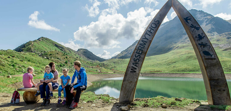 Eine Familie sitzt auf einer Bank am Ufer des Wassermythos Ochsenlacke im Skizentrum St. Jakob i. D..  Im Hintergrund sieht man die dazugehörigen Spielgeräte und das umliegende Bergpanorama, welches durch das schöne Wetter gut zur Geltung kommt.
