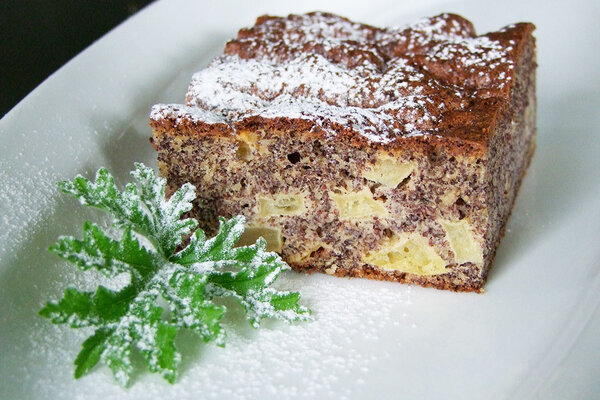 Ein Apfel-Mohnkuchen auf einem Teller, mit einem grünen Blatt und Puderzucker dekoriert.