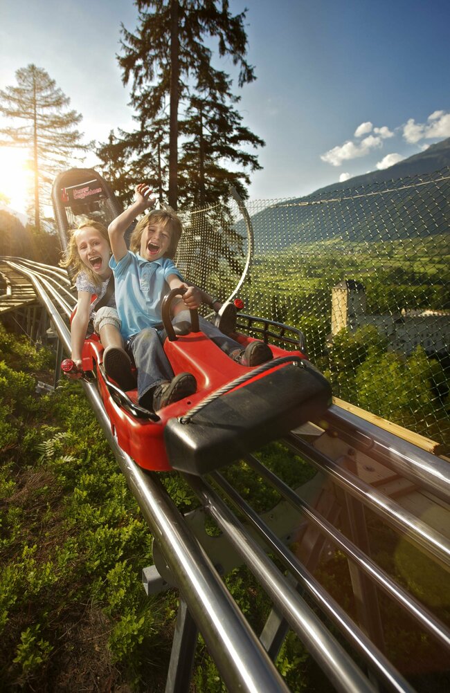Ganzjahresrodelbahn Osttirodler am Hochstein Zwei Kinder fahren lachend mit der roten Sommerrodelbahn in Lienz in Osttirol. Im Hintergrund steht Schloss Bruck.
