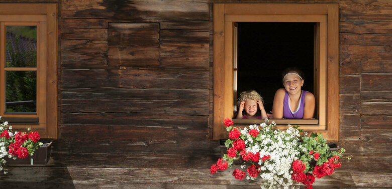 Zwei Kinder schauen aus dem Fenster eines Bauernhofs. An den Balkonen sind weiße und rote Blumen.