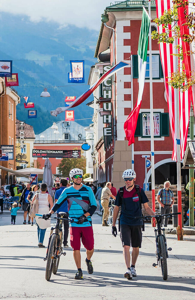 Zwei Radfahrer schieben ihre Fahrräder durch die mit Fahnen geschmückte Lienzer Innenstadt.