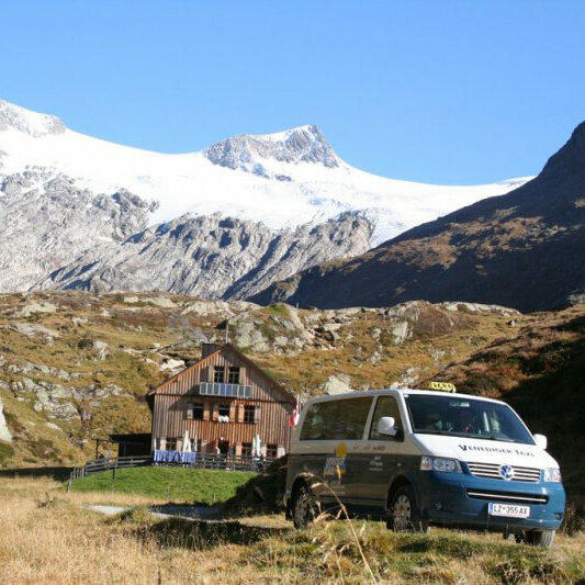 Berge Ein blau-weißer Taxi-Bus vor einem Holzhaus mit schneebedeckten Bergen im Hintergrund.