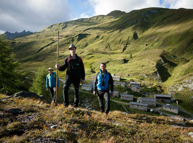 Rangertour Drei Erwachsene wandern durch Umgebung der Jagdhausalmen. Im Hintergrund stehen mehrere alte Gebäude der Jagdhausalmen.
