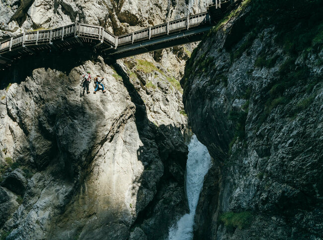 Zwei Personen auf dem Klettersteig in der Galitzenklamm. Sie befinden sich unterhalb des hölzernen Weges und neben einem Wasserfall.