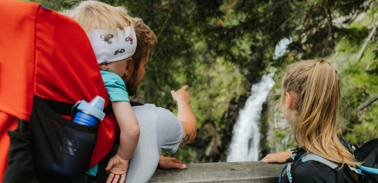 Ein Mann mit einem kleinen Kind auf dem Rücken zeigt einem Mädchen den Wasserfall im Kristeinertal.