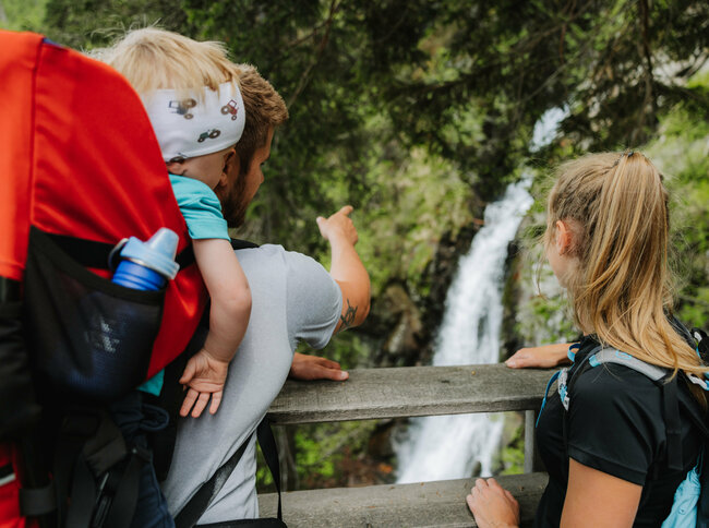Wasserfall Familienwanderung Kristeinertal Ein Mann mit einem kleinen Kind auf dem Rücken zeigt einem Mädchen den Wasserfall im Kristeinertal.