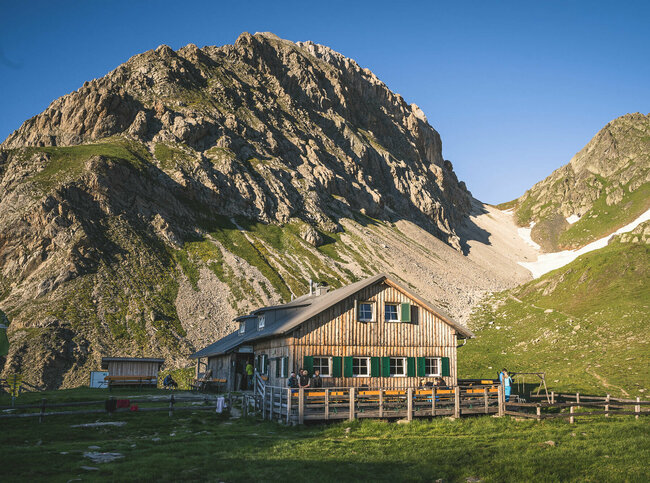 Ein rustikales Berghaus aus Holz mit grünen Fensterläden steht in einer sonnigen, alpinen Wiesenlandschaft. Im Hintergrund ragen steile, felsige Berggipfel auf, teils mit Schneefeldern bedeckt. Vor dem Haus sitzen und stehen einige Menschen, eine Holzterrasse mit Bänken lädt zum Verweilen ein
