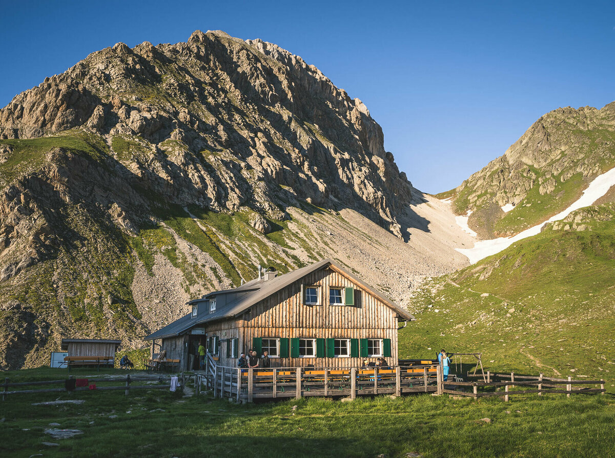 Obstanser Seehütte Ein rustikales Berghaus aus Holz mit grünen Fensterläden steht in einer sonnigen, alpinen Wiesenlandschaft. Im Hintergrund ragen steile, felsige Berggipfel auf, teils mit Schneefeldern bedeckt. Vor dem Haus sitzen und stehen einige Menschen, eine Holzterrasse mit Bänken lädt zum Verweilen ein