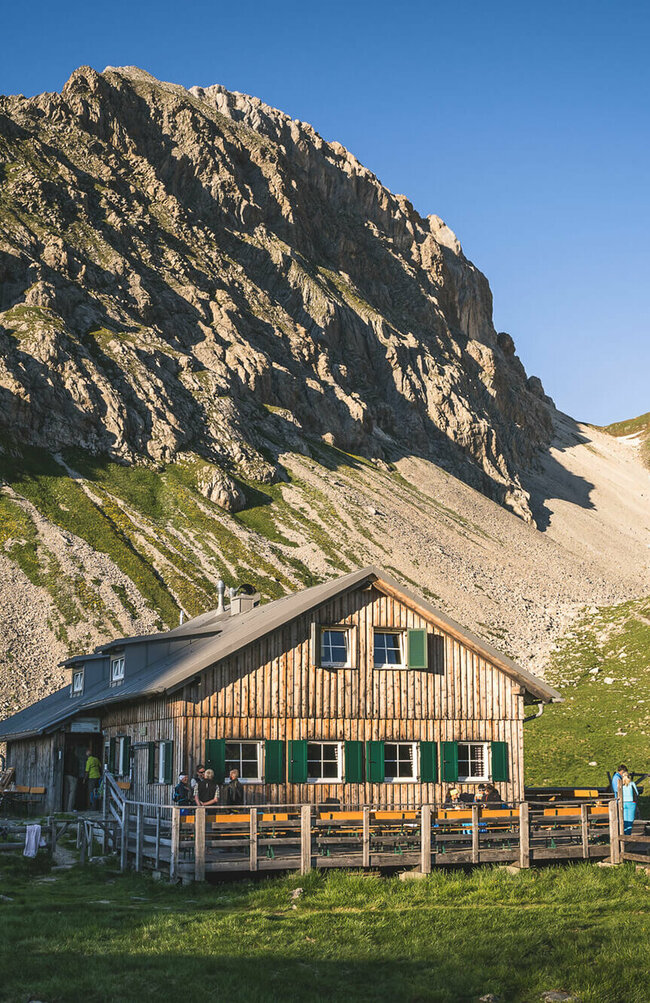 Obstanser Seehütte Ein rustikales Berghaus aus Holz mit grünen Fensterläden steht in einer sonnigen, alpinen Wiesenlandschaft. Im Hintergrund ragen steile, felsige Berggipfel auf, teils mit Schneefeldern bedeckt. Vor dem Haus sitzen und stehen einige Menschen, eine Holzterrasse mit Bänken lädt zum Verweilen ein