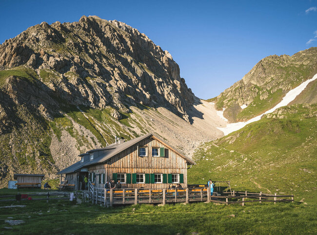 Ein rustikales Berghaus aus Holz mit grünen Fensterläden steht in einer sonnigen, alpinen Wiesenlandschaft. Im Hintergrund ragen steile, felsige Berggipfel auf, teils mit Schneefeldern bedeckt. Vor dem Haus sitzen und stehen einige Menschen, eine Holzterrasse mit Bänken lädt zum Verweilen ein