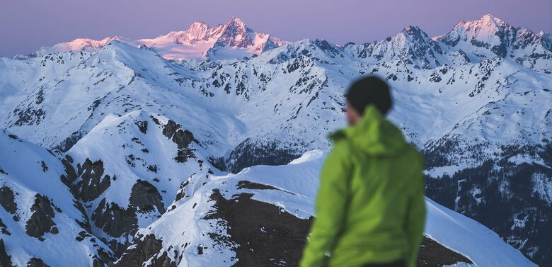 Die höchsten Berggipfel im Hintergrund genießen die letzten Sonnenstrahlen und erstraheln in einem zarten, rötlich gefärbten Licht. Ein Skitourengeher im Vordergrund mit grüner Sportjacke beobachtet dieses zauberhafte Schauspiel.