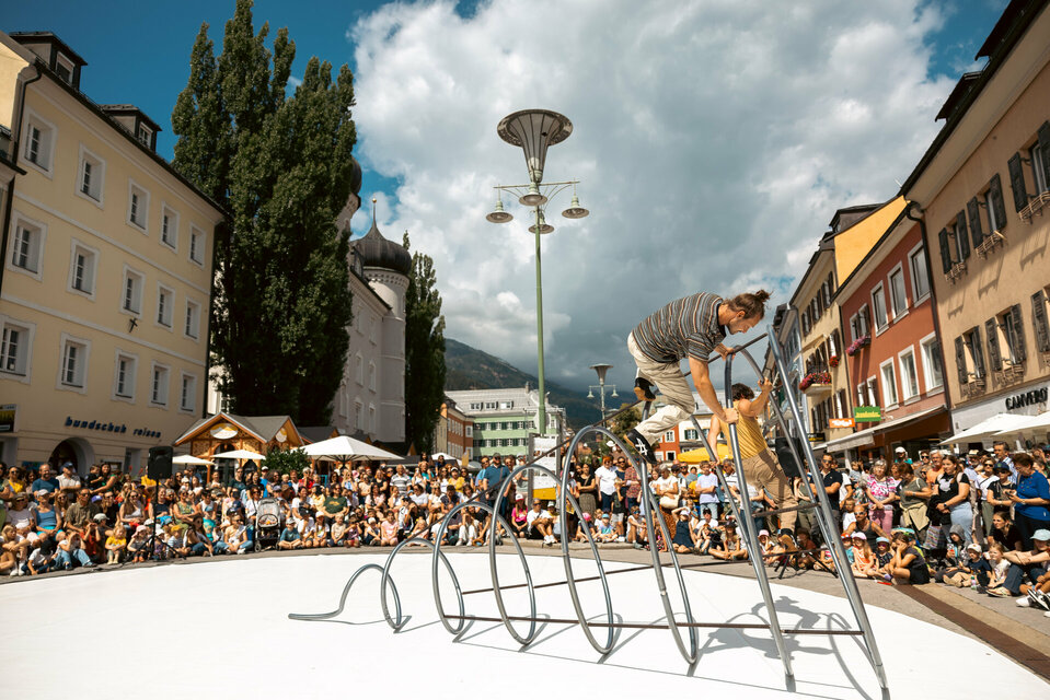 Zwei Akrobaten von The Tea Time Company turnen im Rahmen von Olala auf einer großen Spirale aus Eisen im Kreis am Lienzer Hauptplatz. Viele Schaulustige sind hier am Lienzer Hauptplatz, um sich das Spektakel anzusehen.