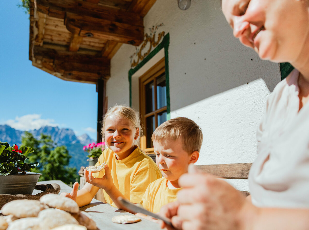 Kinderfreundliche Unterkunft in Nußdorf-Debant Eine Frau und zwei Kinder mit gelben T-Shirts sitzen vor einem Bauernhaus auf einer Bank und verköstigen frisches Brot.