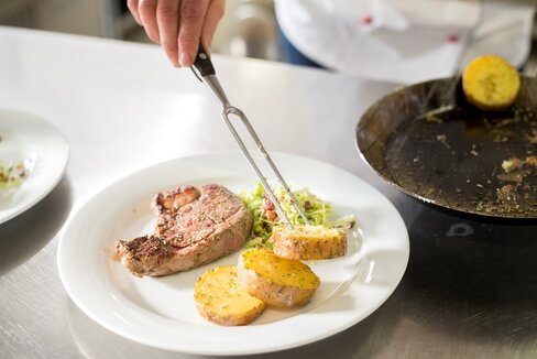 Anrichten von einem Stück Fleisch und dicke Kartoffelscheiben mit Krautsalat auf einem weißen Teller auf demLenzingerhof.
