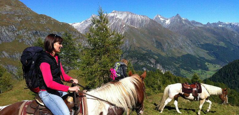 Zwei Frauen reiten auf gescheckten Pferden in einer tollen Bergkulisse. Außerdem läuft ein Pferd ohne Reiter mit Sattelzeug herum.