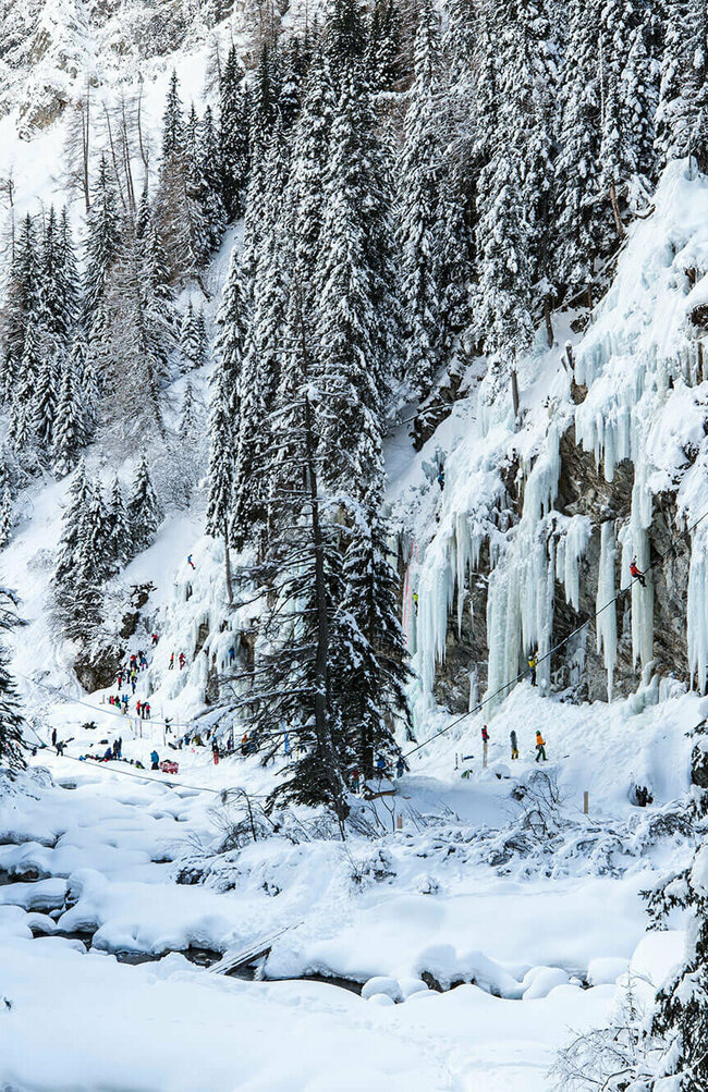 Eiskletterpark Osttirol Drei Eiskletterer stehen vor der Eiswand und sehen auf ihr nach oben. Umrahmt wird der Eiskletterpark von tief verschneiten dunkelgrünen Tannenbäumen.