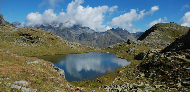 Einer der Neualpenseen am Lienzer Zettersfeld, im Vordergrund der klare Bergsee, im Hintergrund ein mächtiges Bergmassiv unter blauem Himmel.