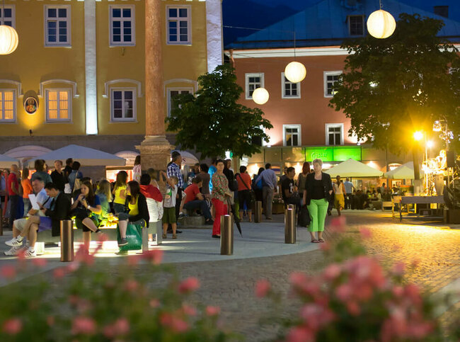 Aufnahme von mehreren Personen beim Moonlight Shoppen in Lienz. Es ist Nacht, die Straßenbeleuchtung ist eingeschalten und das Foto wurde vor einem Blumenbeet gemacht.