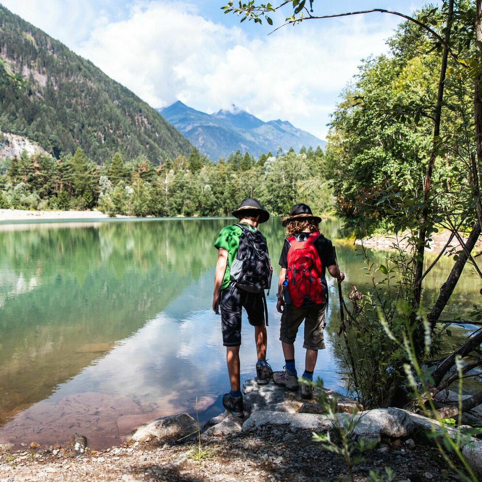 Seeblick Zwei Personen mit Wanderausrüstung und Hüten stehen an einem See und blicken in die Ferne.