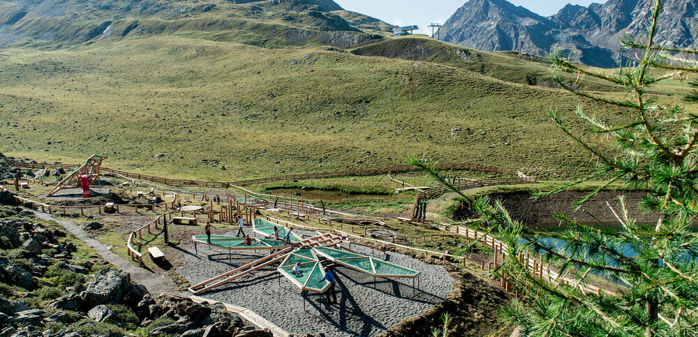 Wassermythos Ochsenlacke St. Jakob Brunnalm Der Blick auf den Wassermythos Ochsenlacke im Skizentrum St. Jakob i. D.. Man kann neben einem Stück des Ufers der Ochsenlacke auch zwei Spielgeräte erkennen, auf denen gerade Kinder klettern. Die Sonne lässt die Umgebung und die umliegende Bergwelt in einem warmen Licht erstrahlen.