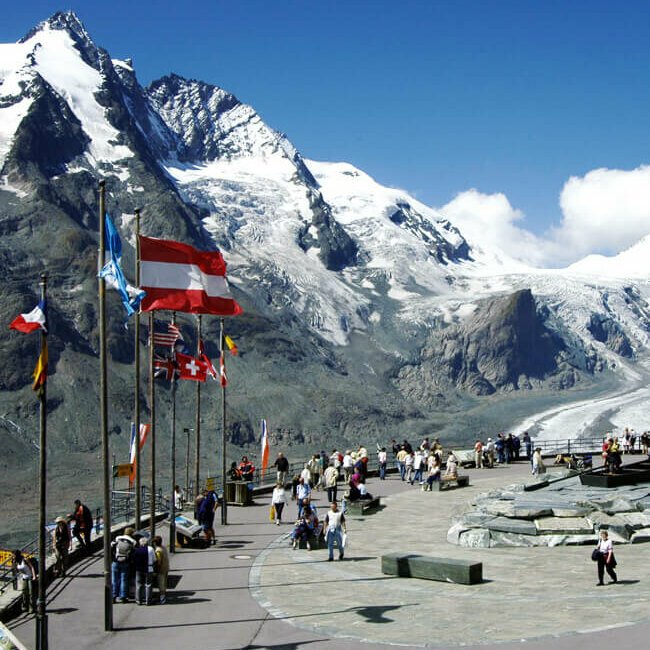 Glockner Weitblick von der Franz-Josefs-Höhe: Parkplatz mit wehenden Fahnen, dahinter die mächtige Pasterze und der imposante, schneebedeckte Großglockner.