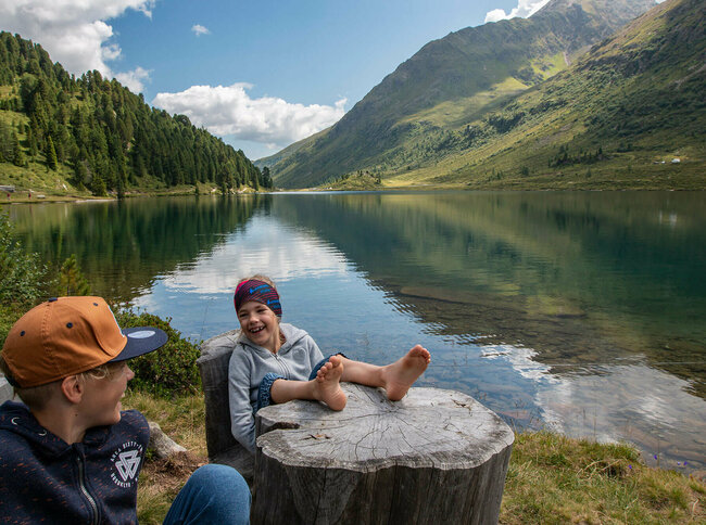Obersee am Staller Sattel Zwei lachende Buben, die am Rand eines Gebirgssees sitzen, wobei einer seine bloßfüßigen Beine auf einem glatten, aber verwitterten Baumstumpf liegen lässt. Im Hintergrund spiegelt sich der Horziont im glasklaren Wasser des Gebirgssees.