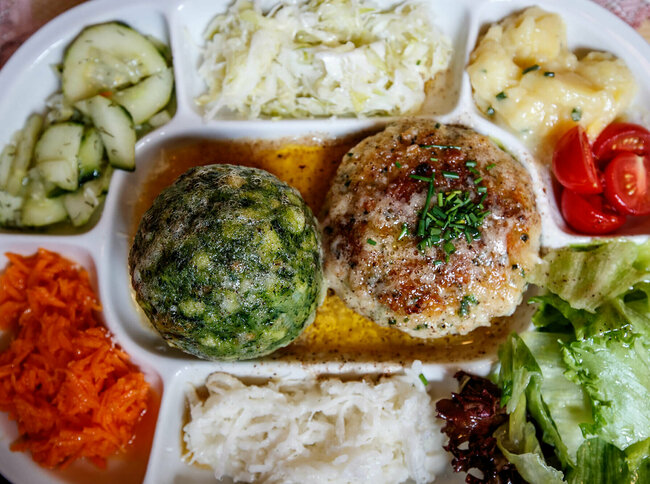Spinatknödel und Kaspressknödel in brauner Butter. Ringsherum verschiedene bunte Salate auf einem Teller angerichet.