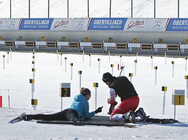 Eine Frau versucht sich am Schießstand im Biathlonzentrum Obertilliach am Schießen. Dabei wird sie von einer rechts neben ihr knieenden Person unterstützt.
