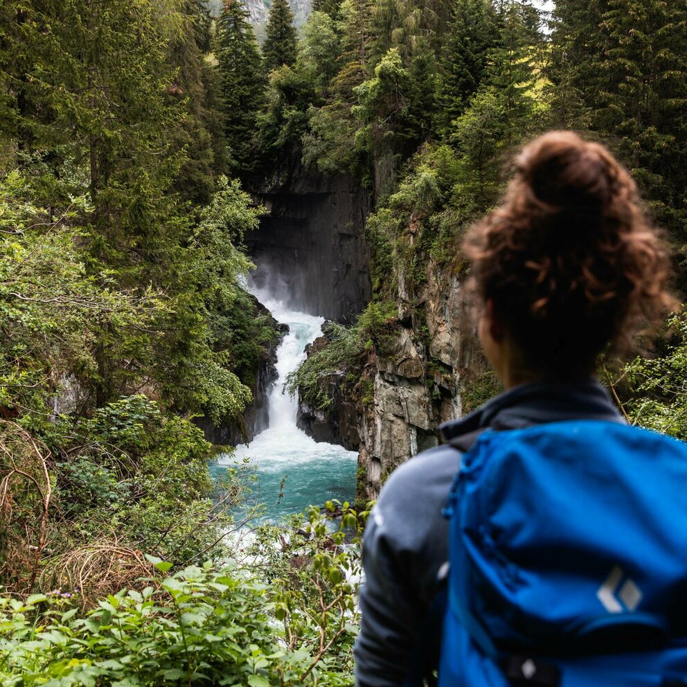 Eine Frau mit blauem Rucksack und braunem Dutt steht vor einem Wasserfall mit Blickrichtung zum Wasser.