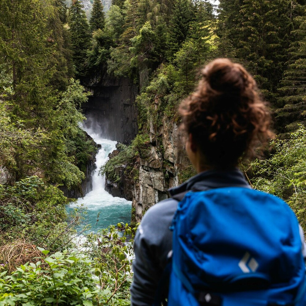 Eine Frau mit blauem Rucksack und braunem Dutt steht vor einem Wasserfall mit Blickrichtung zum Wasser.