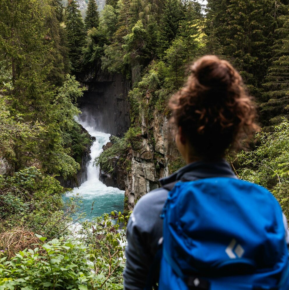 Frau mit Wasserfall Eine Frau mit blauem Rucksack und braunem Dutt steht vor einem Wasserfall mit Blickrichtung zum Wasser.