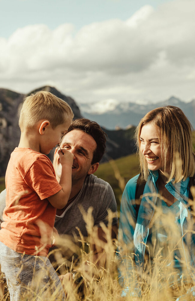 kindermeile-obertilliach Zu sehen ist ein Elternpaar mit ihrem kleinen Sohn, die im hohen Gras entlang der Kindermeile am Golzentipp den Moment zu dritt genießen.