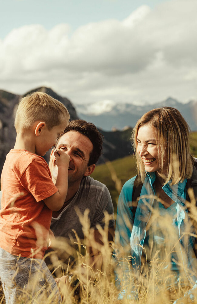 kindermeile-obertilliach Zu sehen ist ein Elternpaar mit ihrem kleinen Sohn, die im hohen Gras entlang der Kindermeile am Golzentipp den Moment zu dritt genießen.