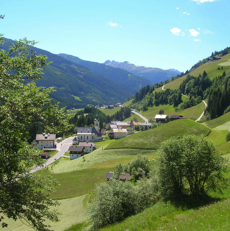 Blick von einem höher gelegenen Hang auf die Pfarrkirche in Untertilliach. Auf den umliegenden saftig grünen Wiesen finden Mäharbeiten bei strahlend blauem Himmel statt.