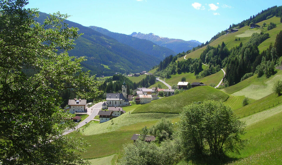 Untertilliach Blick von einem höher gelegenen Hang auf die Pfarrkirche in Untertilliach. Auf den umliegenden saftig grünen Wiesen finden Mäharbeiten bei strahlend blauem Himmel statt.