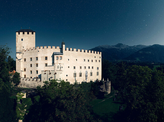 Aussenaufnahme bei Nacht von Schloss Bruck in Lienz mit Bergkulisse und Sternenhimmel im Hintergrund.