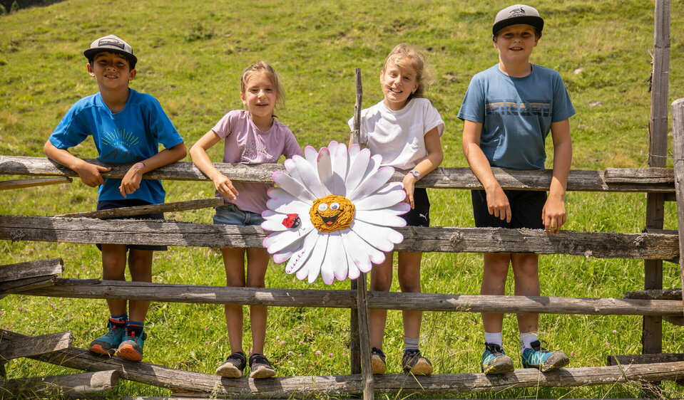 Die vier Kinder stehen mit Gisela Gänseblümchen in ihrer Mitte auf einem Holzzaun vor einer grünen Wiese.