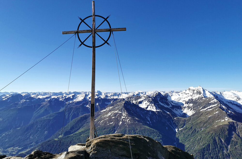 Gipfelkreuz am vorderen Sajatkopf mit frühlingshaften Bergpanorama im Hintergrund.