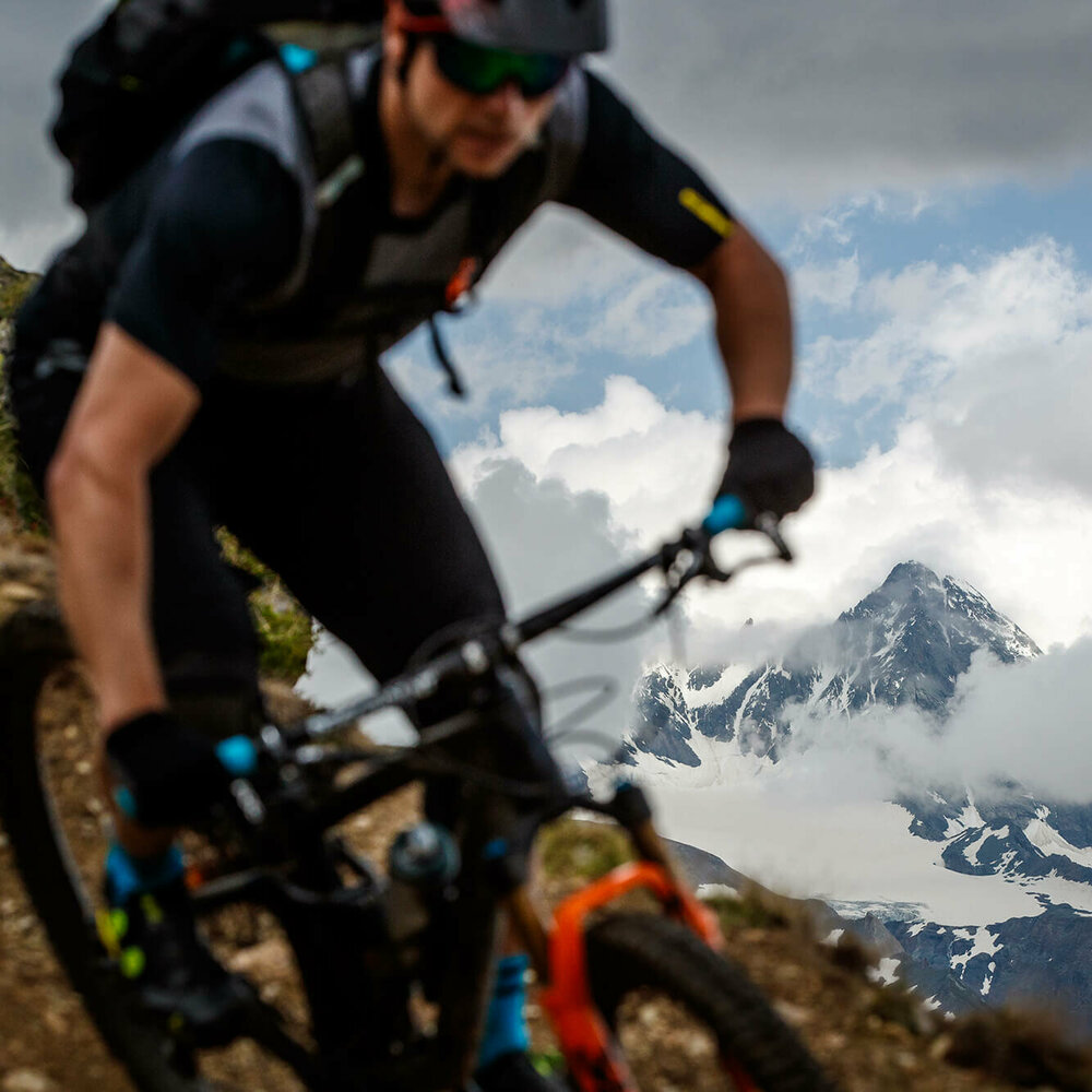 Bikepark Grossglockner Resort Abwärts fahrender Mountainbiker mit dunkler, kurzärmeliger Bekleidung und schwarzer Hose im Vordergrund, dahinter ein zum Teil vergletscherter Berg, der sich aus dem Nebel befreit.