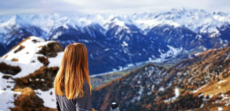 Eine Frau blickt von einem Berg mit einigen Schneefeldern hinunter ins Tal und die gegenüberliegenden, noch komplett schneebedeckten Gipfel.