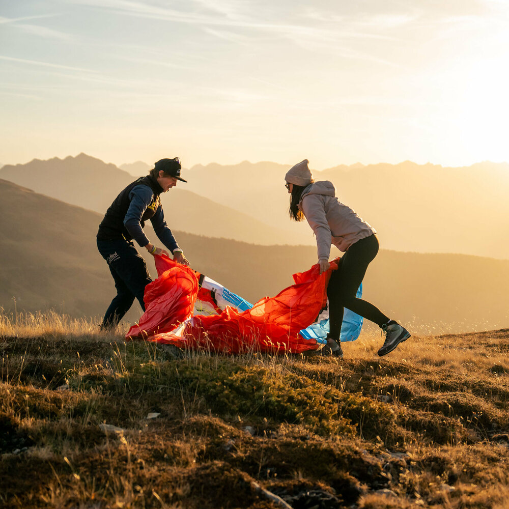 Ein Mann und eine Frau stehen auf einer Bergwiese in Osttirol in der Morgensonne und falten einen roten Gleitschirm zusammen.